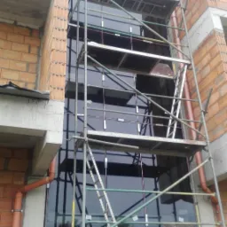 Montaż dużego okna fasadowego na rusztowaniu w trakcie budowy, widoczne cegły i rury spustowe.