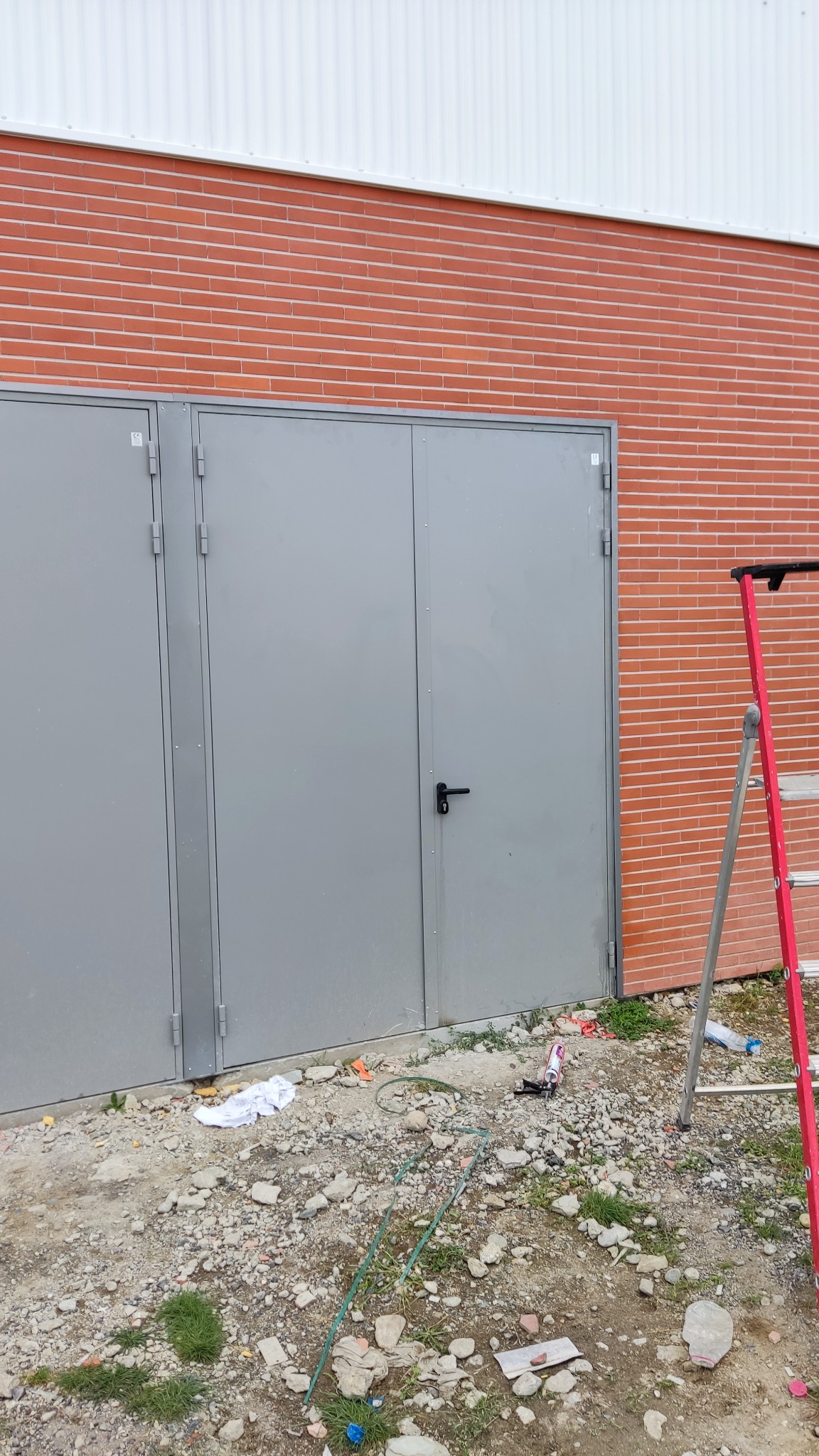 Szare metalowe drzwi przemysłowe z czarną klamką, zamontowane w ścianie z czerwonej cegły, z widoczną czerwoną drabiną i gruzem budowlanym na ziemi.