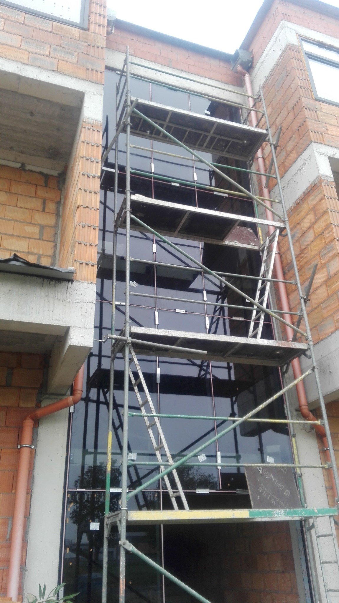 Montaż dużego okna fasadowego na rusztowaniu w trakcie budowy, widoczne cegły i rury spustowe.