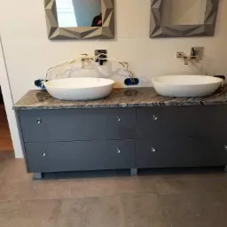 Szara szafka łazienkowa z blatem z szarego kamienia, dwie białe umywalki nablatowe, lustra w geometrycznych ramach i wystające kable instalacji wodnej w trakcie montażu.