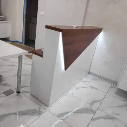 Nowoczesna lada recepcyjna z białym korpusem i drewnianym blatem w kształcie trójkąta, podświetlona od spodu, na tle marmurowej podłogi i ściany z dekoracyjnym tynkiem.