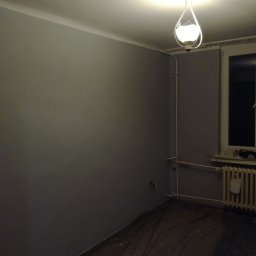Gładzix Hubert Wójcik - Świeżo pomalowane na szaro ściany pokoju z widoczną lampą sufitową w kształcie łzy i zabezpieczoną folią podłogą przed malowaniem.