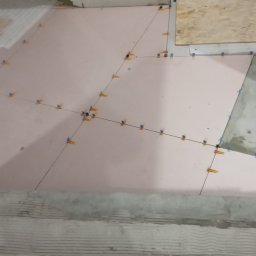 Gładzix Hubert Wójcik - Podłoga w trakcie układania płytek, widoczne krzyżyki dystansowe, czarne linie wyznaczające położenie płytek i fragment schodów w tle.