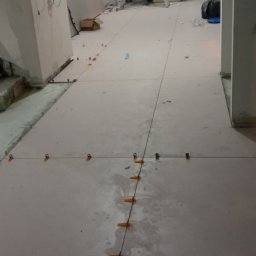 Gładzix Hubert Wójcik - Proces układania jasnych płytek podłogowych w korytarzu, widoczne systemy poziomowania płytek, drabina i instalacje na suficie w tle, osoba przechodząca w oddali.