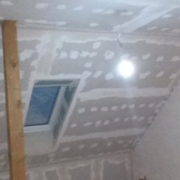 Gładzix Hubert Wójcik - Poddasze w trakcie wykańczania: widoczne płyty gipsowo-kartonowe na skośnym suficie i ścianach, częściowo zaszpachlowane łączenia, okno dachowe, drabina i prowizoryczne oświetlenie.