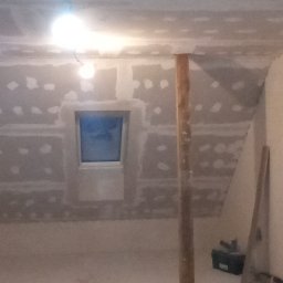 Gładzix Hubert Wójcik - Poddasze w trakcie prac wykończeniowych: zabudowa ścian i sufitu płytami gipsowo-kartonowymi, widoczne szpachlowanie łączeń, okno dachowe, drewniany słup podtrzymujący konstrukcję, surowa...