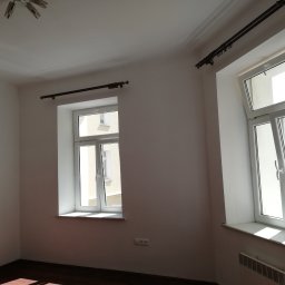 Gładzix Hubert Wójcik - Pomalowane na biało wnętrze pokoju z dwoma oknami z zamontowanymi karniszami, widoczny fragment podłogi i grzejnik.