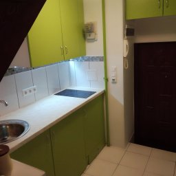 Gładzix Hubert Wójcik - Fragment kuchni z zielonymi szafkami, białym blatem i płytą indukcyjną, widok na mozaikę nad blatem i brązowe drzwi wejściowe.