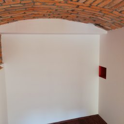 Gładzix Hubert Wójcik - Wnętrze z ceglanym sklepieniem łukowym, białe ściany i ciemna drewniana podłoga, z małym kwadratowym otworem w ścianie pomalowanym na czerwono.