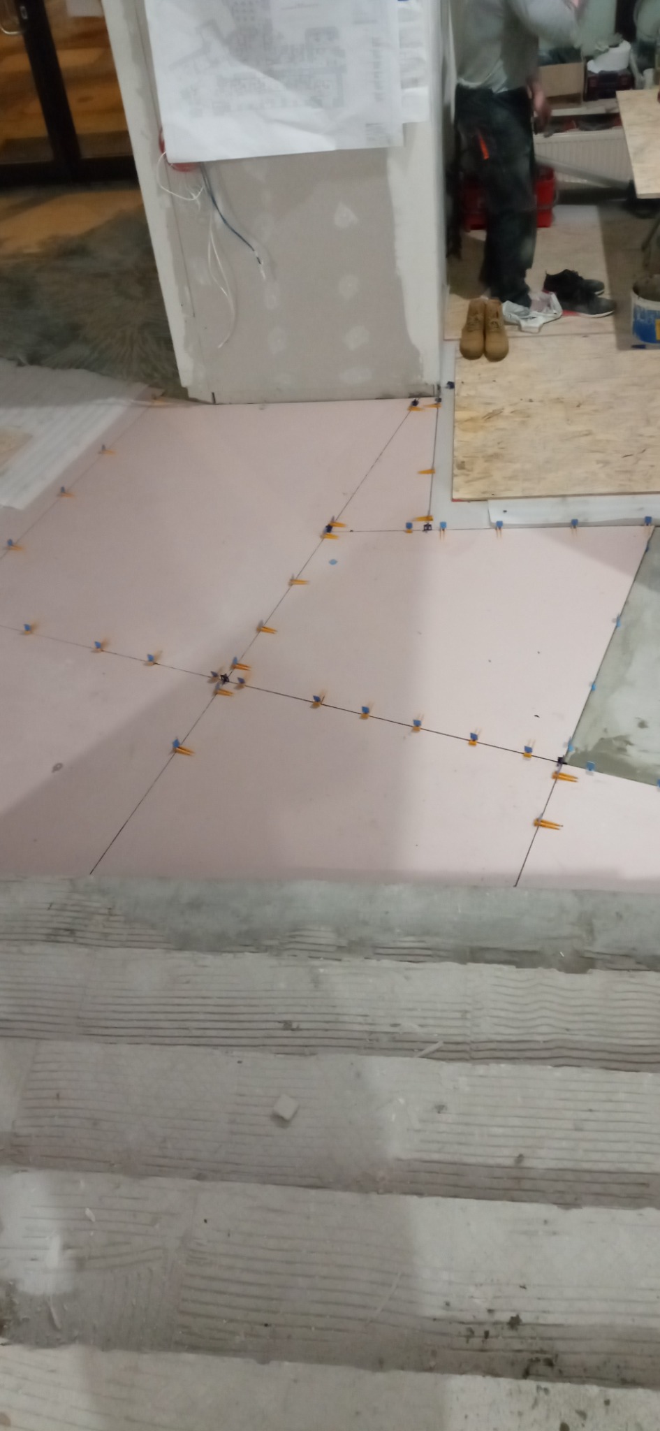 Podłoga w trakcie układania płytek, widoczne krzyżyki dystansowe, czarne linie wyznaczające położenie płytek i fragment schodów w tle.