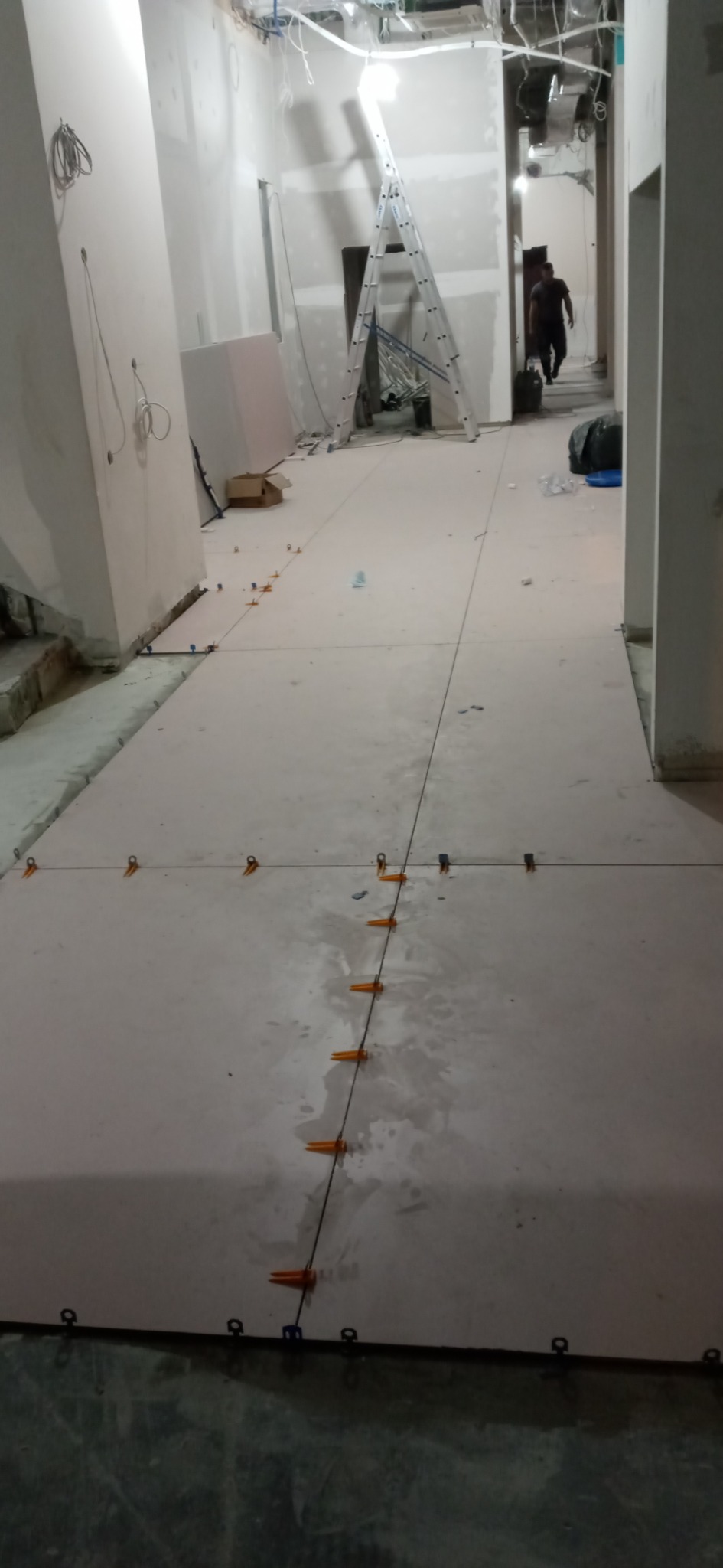 Proces układania jasnych płytek podłogowych w korytarzu, widoczne systemy poziomowania płytek, drabina i instalacje na suficie w tle, osoba przechodząca w oddali.