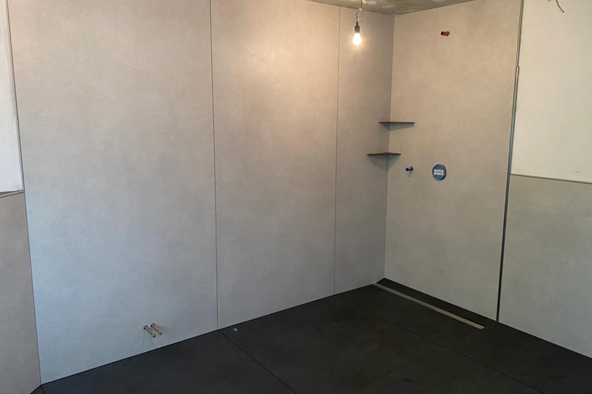 Niedokończona łazienka z szarymi płytami wielkoformatowymi na ścianach, czarną podłogą, odpływem liniowym i prowizorycznym oświetleniem żarówką