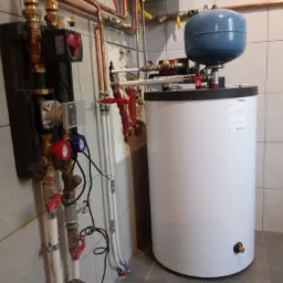 Biały bojler elektryczny z niebieskim zbiornikiem wyrównawczym, podłączony do instalacji z miedzianych rur, zaworów i pomp obiegowych, na tle ściany wyłożonej szarą płytką.