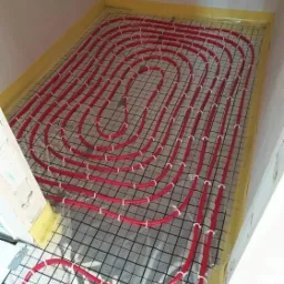 Czerwone rury ogrzewania podłogowego ułożone na czarnej siatce w pomieszczeniu z żółtą folią brzegową, widok z góry.