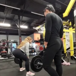 Kobieta podnosi sztangę pod okiem trenera w siłowni, widoczne obciążenia i sprzęt do ćwiczeń.