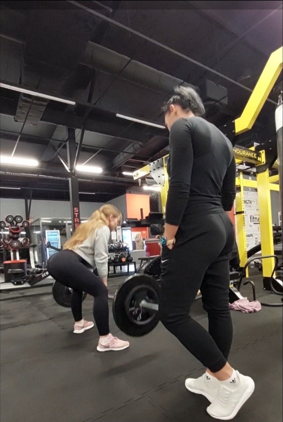 Kobieta podnosi sztangę pod okiem trenera w siłowni, widoczne obciążenia i sprzęt do ćwiczeń.