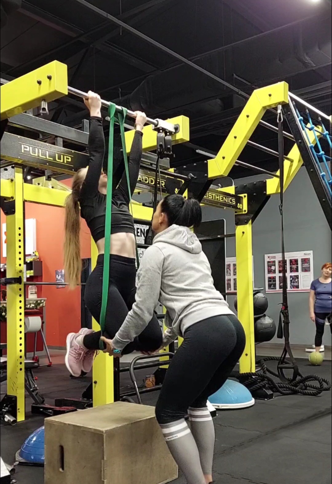 Kobieta wykonuje podciąganie na drążku z pomocą gumy i asystą drugiej kobiety w siłowni, widoczne elementy wyposażenia siłowni w kolorach żółtym i czarnym.
