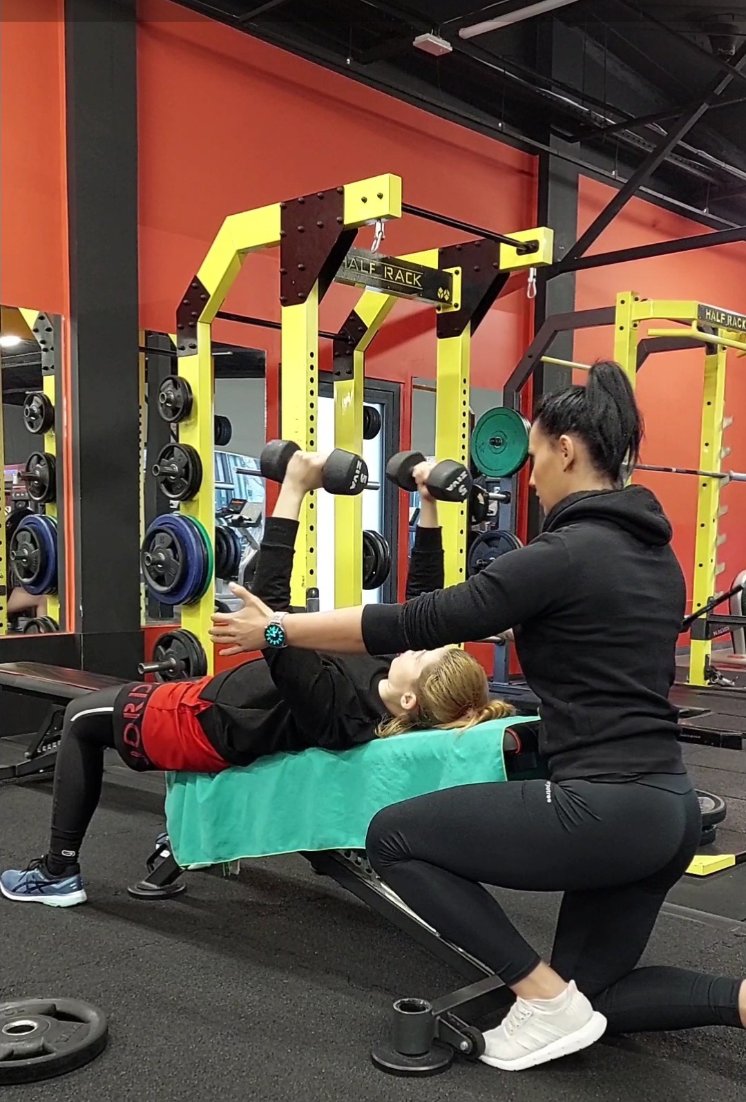 Kobieta ćwiczy wyciskanie hantli leżąc na ławce pod okiem instruktorki w czarnym stroju sportowym w siłowni z żółto-czarną konstrukcją do ćwiczeń.