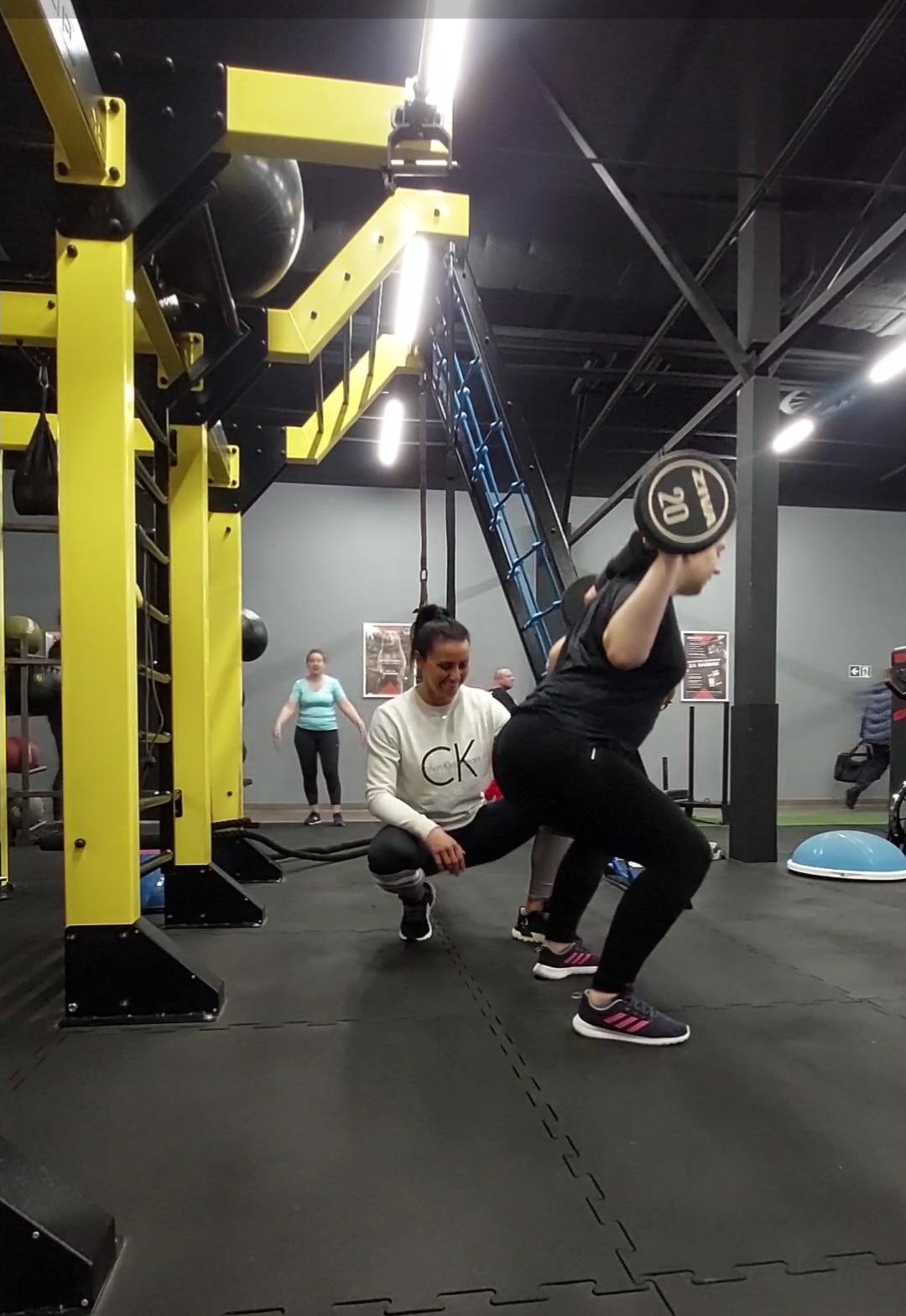 Kobieta wykonuje przysiad z obciążeniem 20 kg pod nadzorem trenera personalnego w siłowni z żółtą konstrukcją do ćwiczeń.