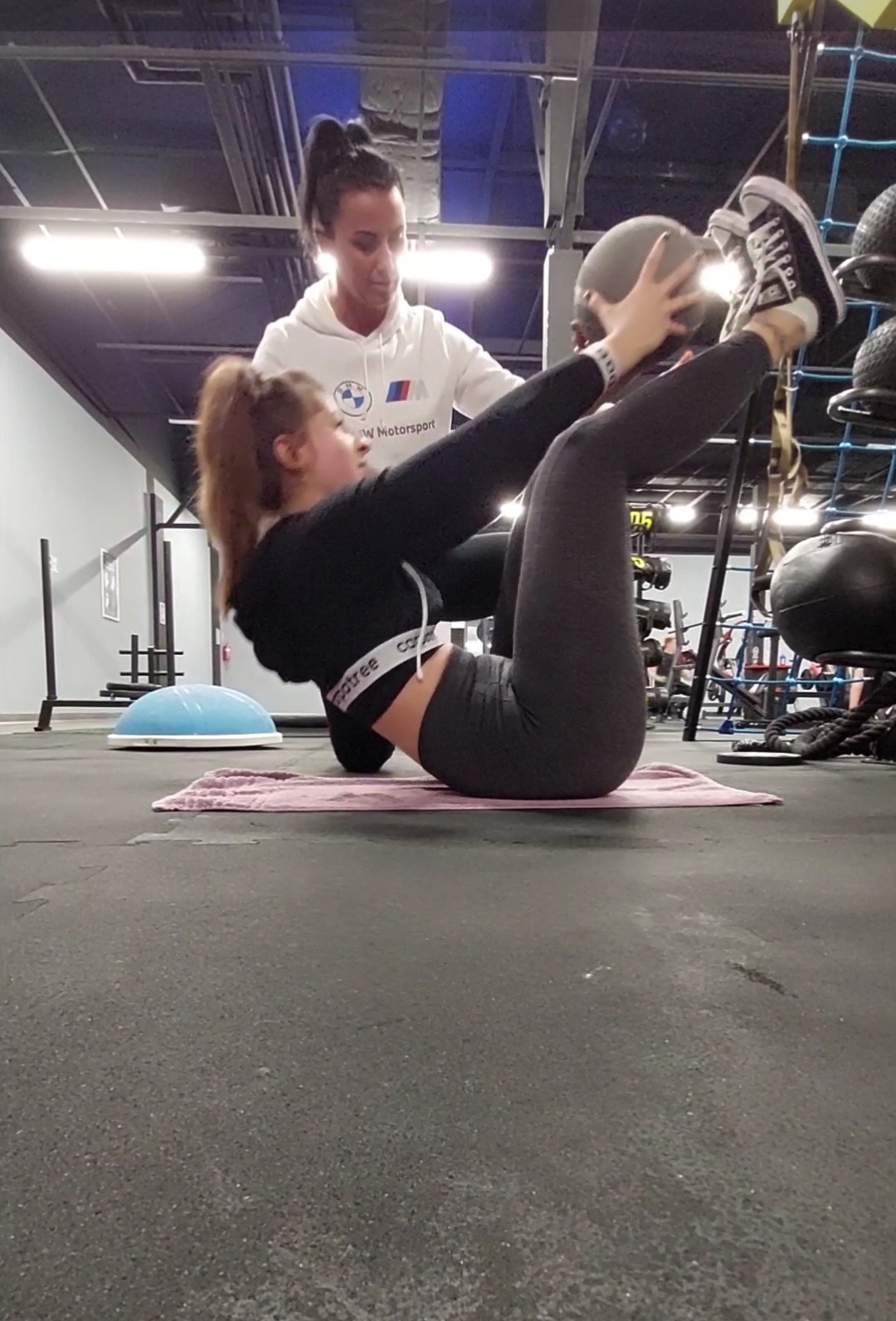 Kobieta wykonuje ćwiczenia brzuszków z piłką lekarską pod nadzorem drugiej kobiety w siłowni. Ćwicząca ma na sobie czarne legginsy i bluzę z krótkim rękawem, a nadzorująca białą bluzę z kapturem...