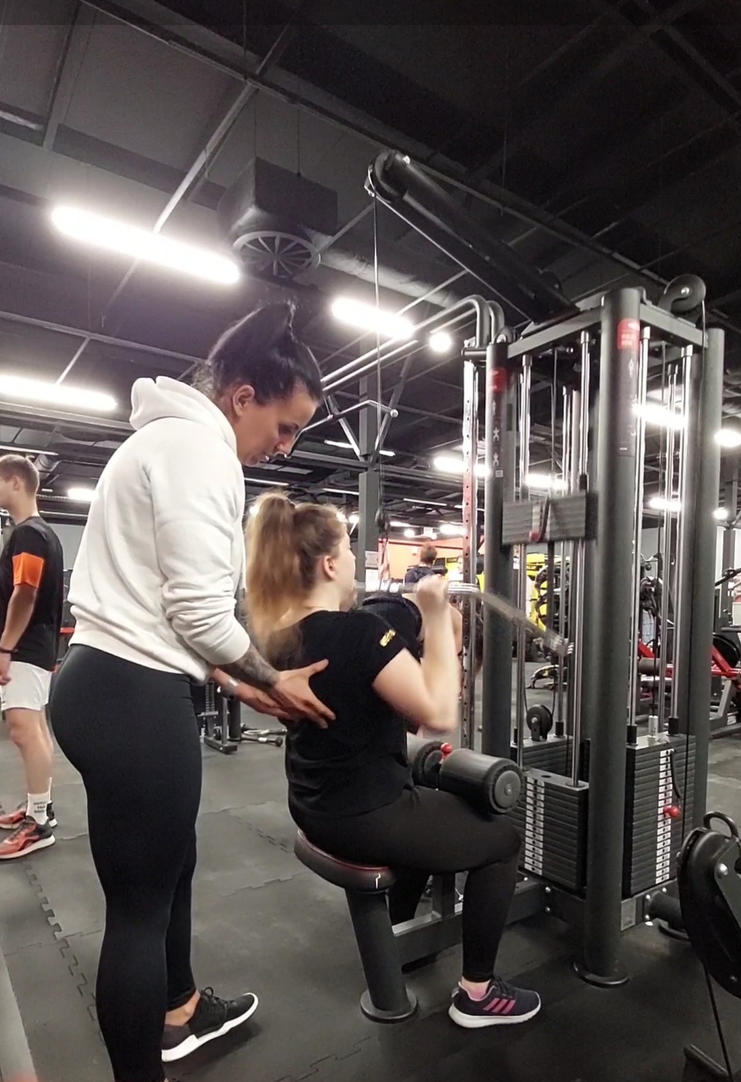 Kobieta ćwiczy na maszynie pod okiem instruktorki w siłowni, instruktorka z tatuażem na ramieniu koryguje postawę ćwiczącej.