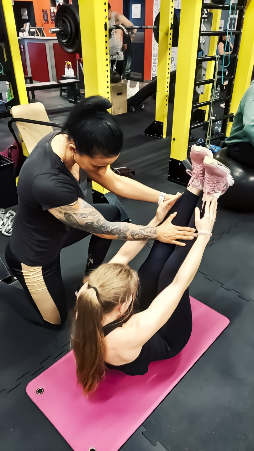 Kobieta z tatuażem na ramieniu pomaga drugiej osobie w ćwiczeniu unoszenia nóg w siłowni na różowej macie do ćwiczeń.
