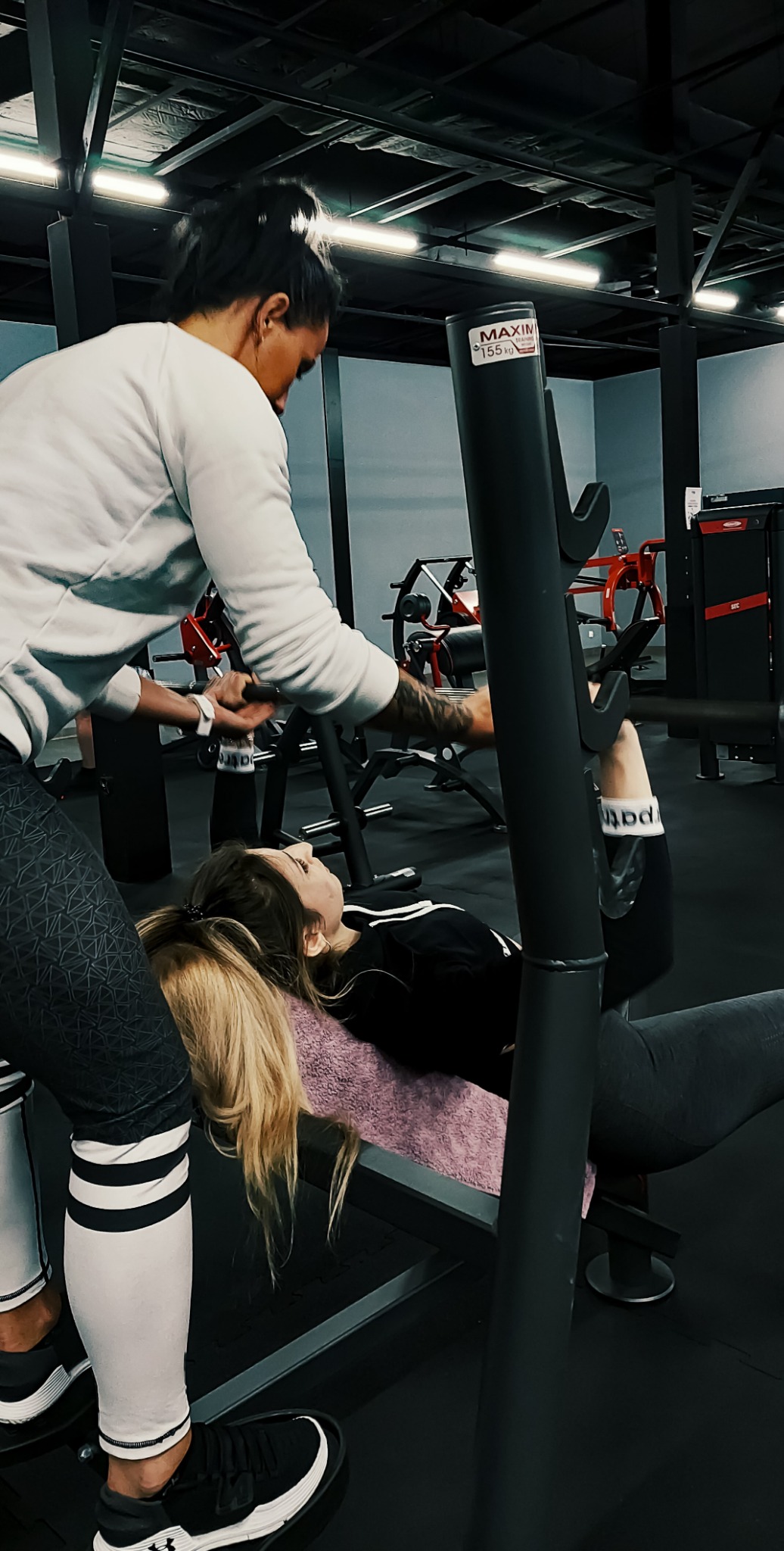 Kobieta ćwiczy wyciskanie sztangi na ławce pod okiem trenera w siłowni, trener asekuruje zza głowy, widoczne obciążenie na sztandze.