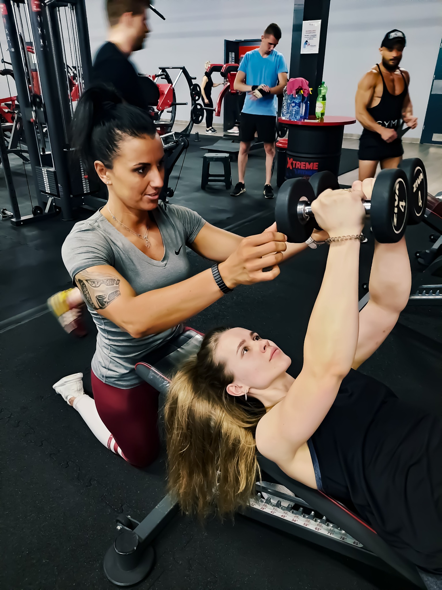 Kobieta z tatuażem na ramieniu pomaga młodej kobiecie ćwiczyć wyciskanie hantli na ławce w siłowni, w tle widoczni inni ćwiczący.