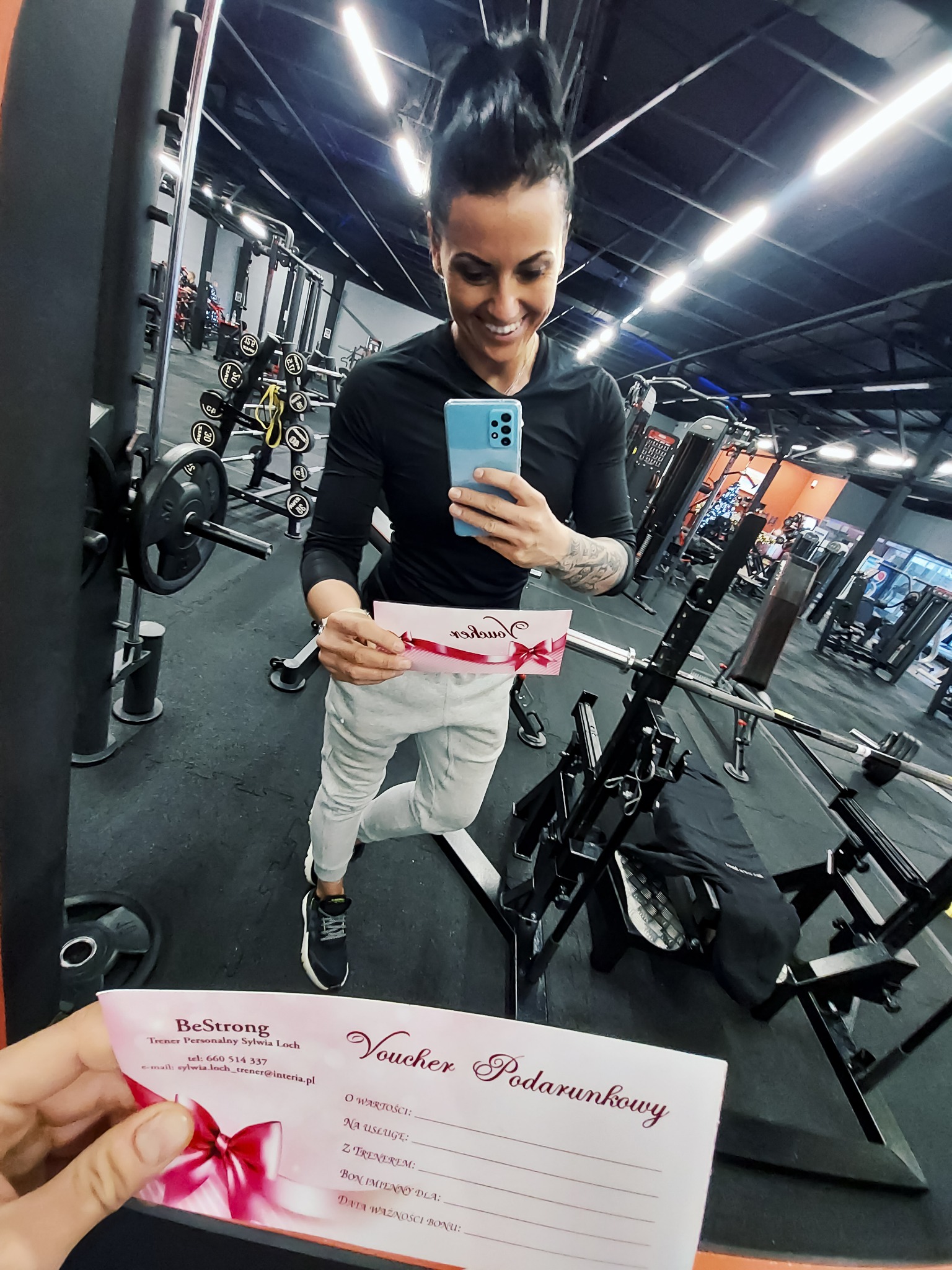 Uśmiechnięta kobieta robi selfie w siłowni, trzymając voucher podarunkowy z logo 'BeStrong Trener Personalny Sylwia Loch', w tle widoczne maszyny do ćwiczeń.