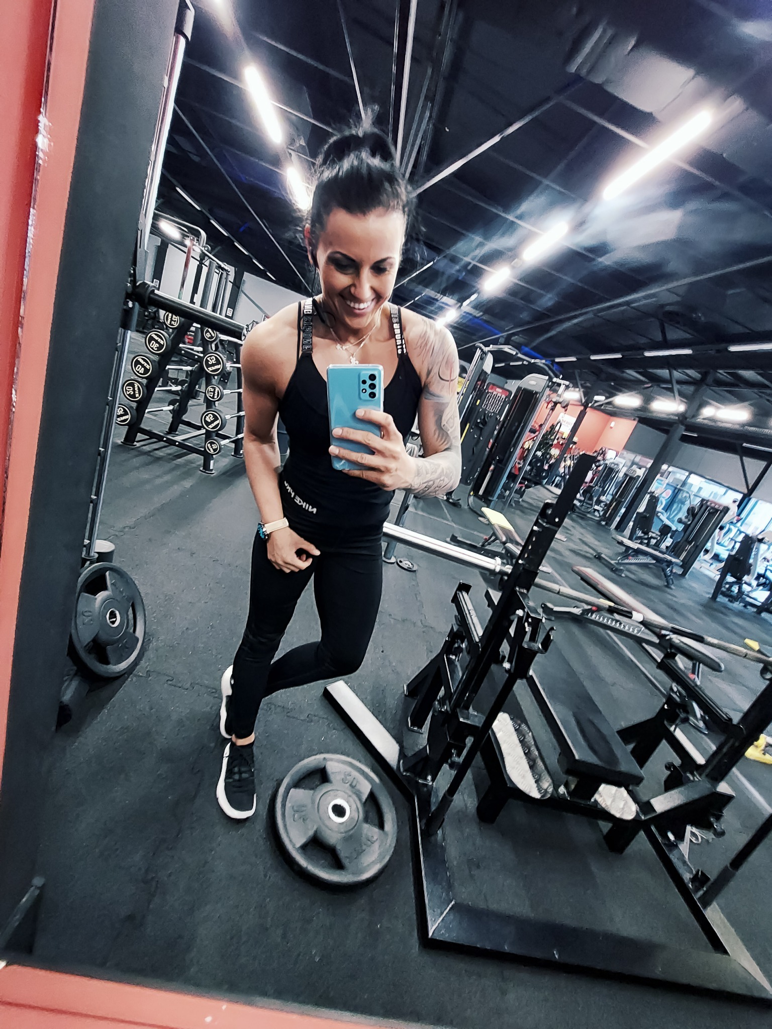 Kobieta w czarnym stroju sportowym robi selfie w lustrze na siłowni, widoczne maszyny do ćwiczeń i obciążenia.