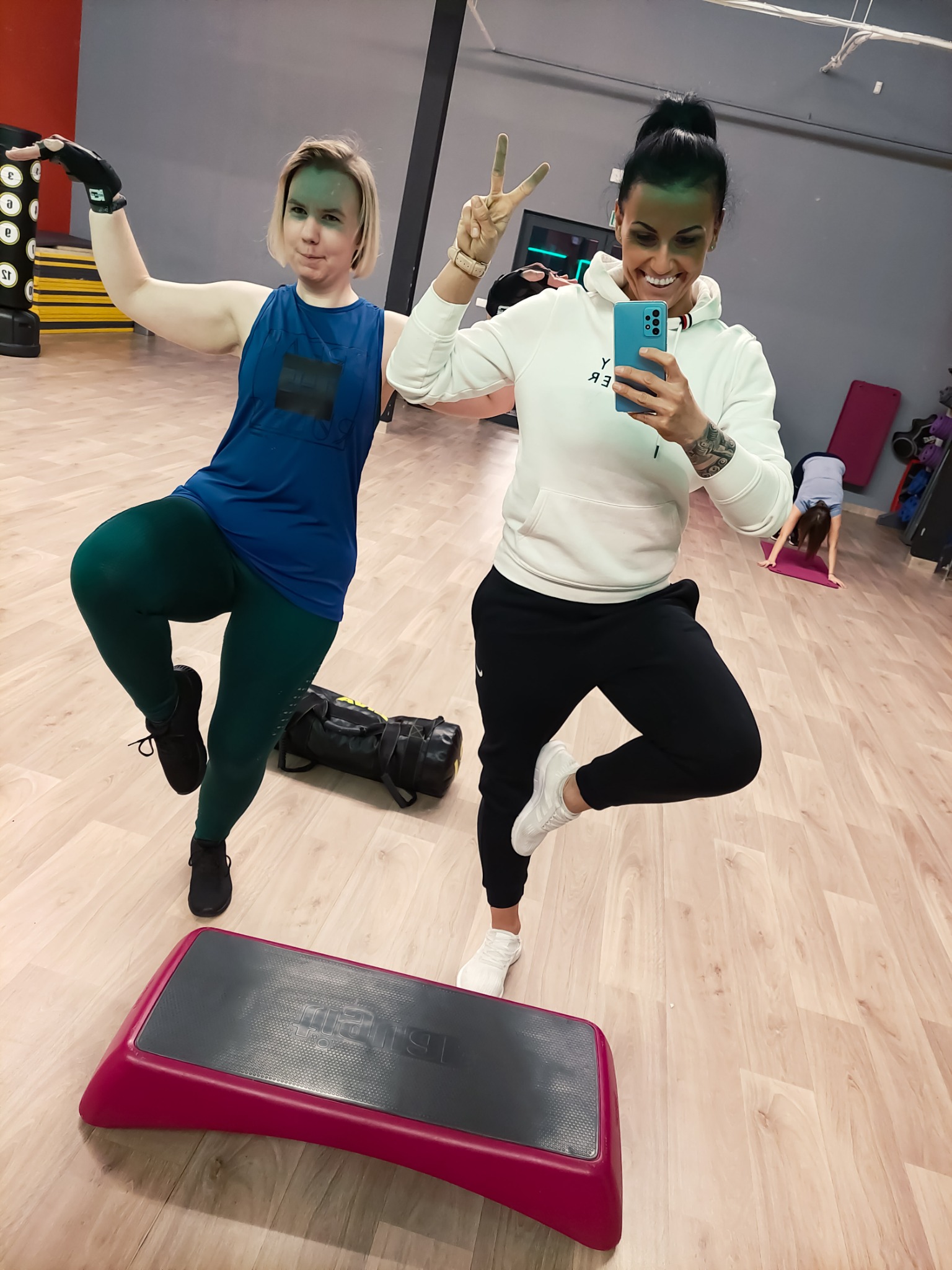 Dwie uśmiechnięte kobiety pozują w sali fitness, jedna robi selfie telefonem, druga stoi na jednej nodze z uniesioną ręką, w tle step, worek treningowy i osoba ćwicząca jogę.