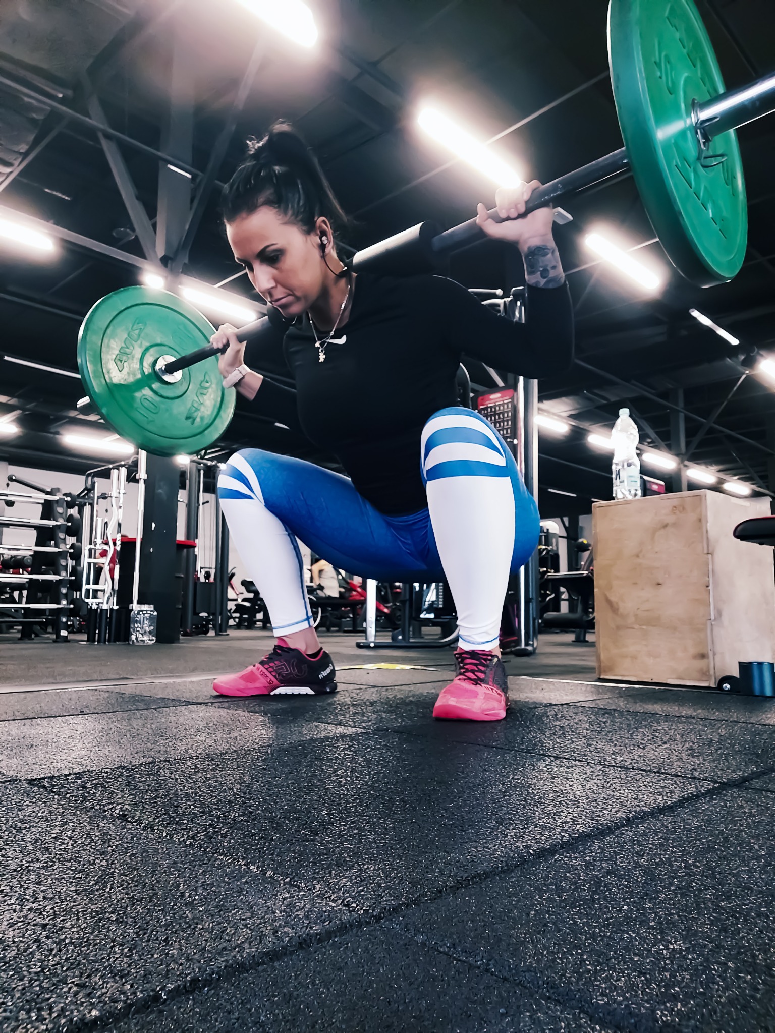 Kobieta w sportowym stroju wykonuje przysiad ze sztangą w siłowni w Częstochowie, widoczna butelka wody i skrzynia do ćwiczeń w tle.