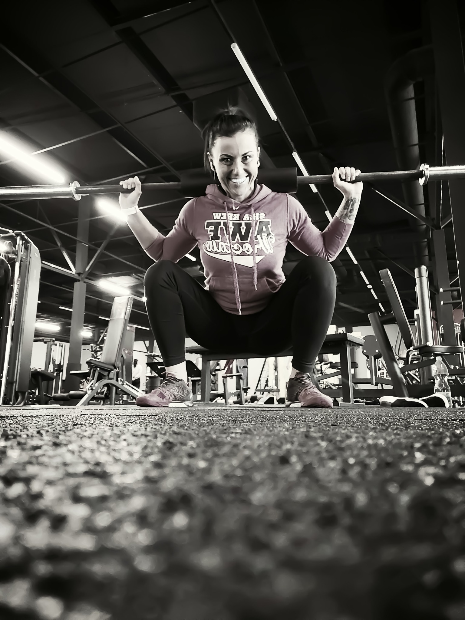 Kobieta w różowej bluzie z kapturem i czarnych legginsach wykonuje przysiad ze sztangą na siłowni, uśmiechając się do kamery, z widocznym tatuażem na lewym przedramieniu i butami sportowymi.