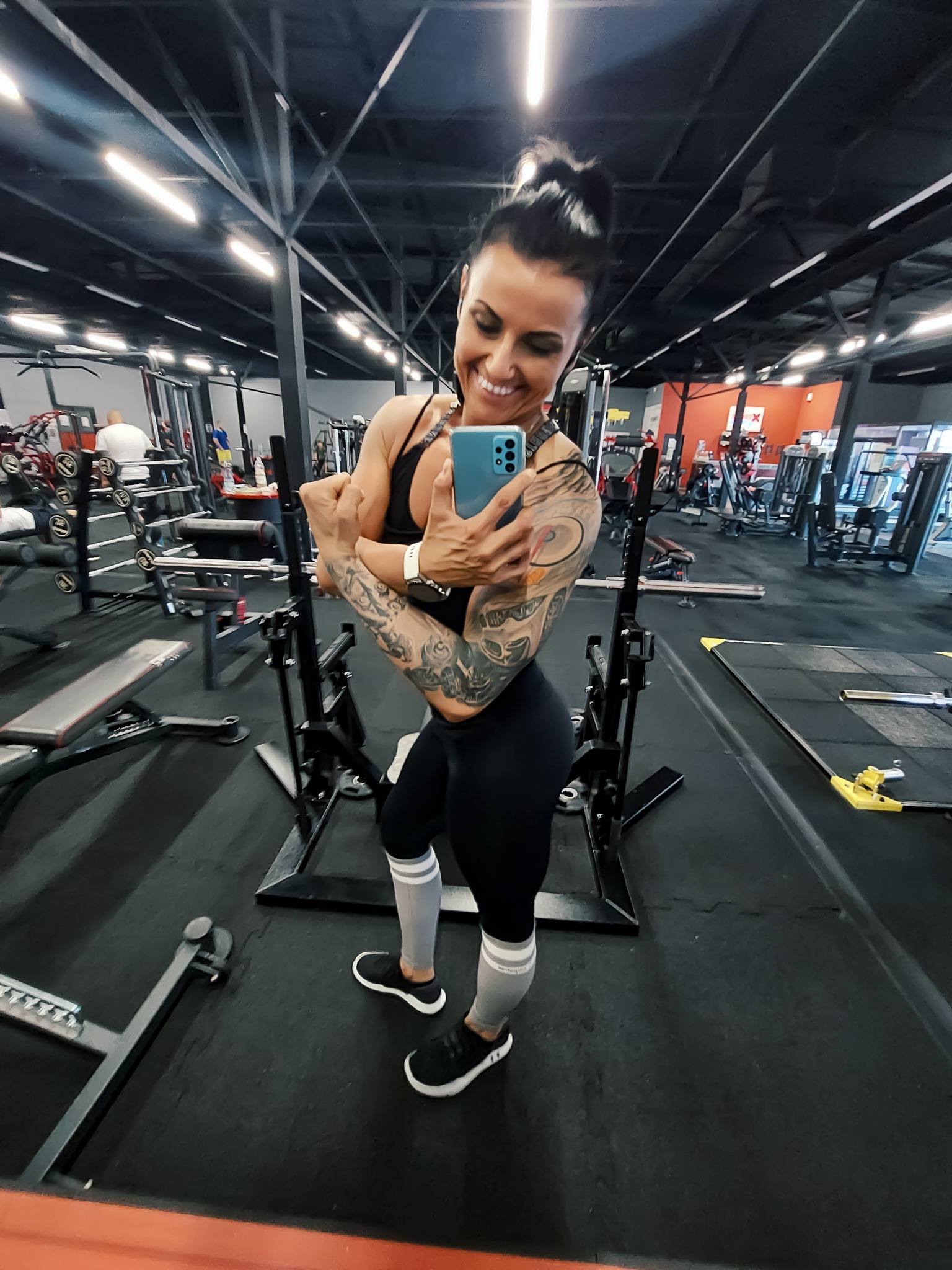 Uśmiechnięta kobieta w sportowym stroju, robi selfie w lustrze na siłowni, prezentując wytatuowane ramiona.