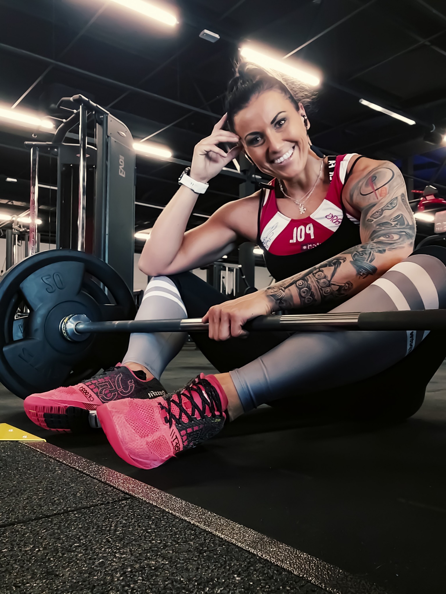 Uśmiechnięta kobieta z tatuażami w sportowym stroju siedzi na podłodze w siłowni, trzymając sztangę obciążoną talerzami 50 kg, w różowych butach sportowych.