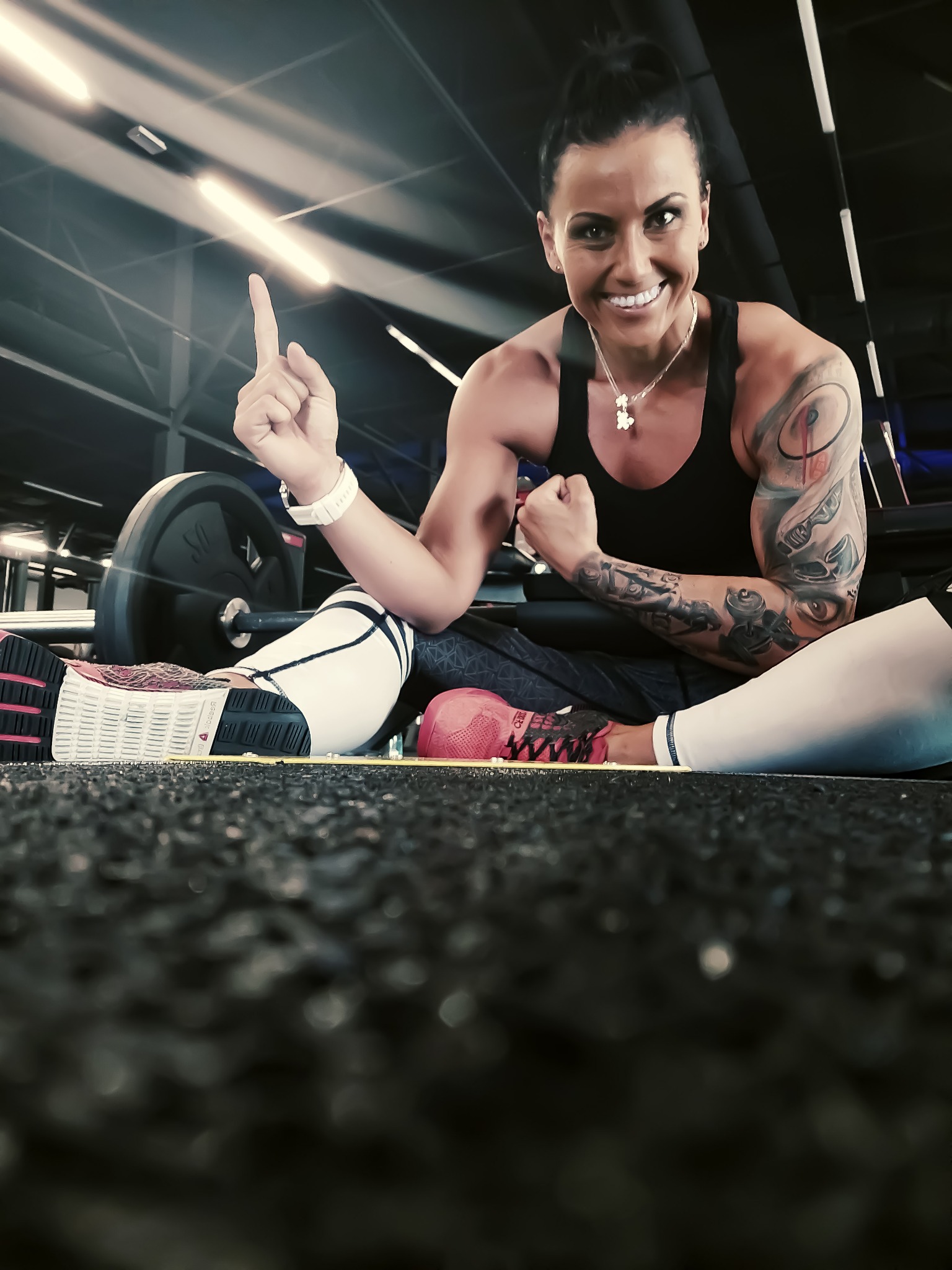Uśmiechnięta kobieta z tatuażami, siedząca na podłodze w siłowni, pokazująca palcem do góry, obok sztangi i hantli.