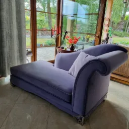 Elegancki, fioletowy szezlong z welurową tapicerką, ozdobnymi nóżkami i poduszką, ustawiony w jasnym pokoju z dużymi oknami wychodzącymi na ogród.