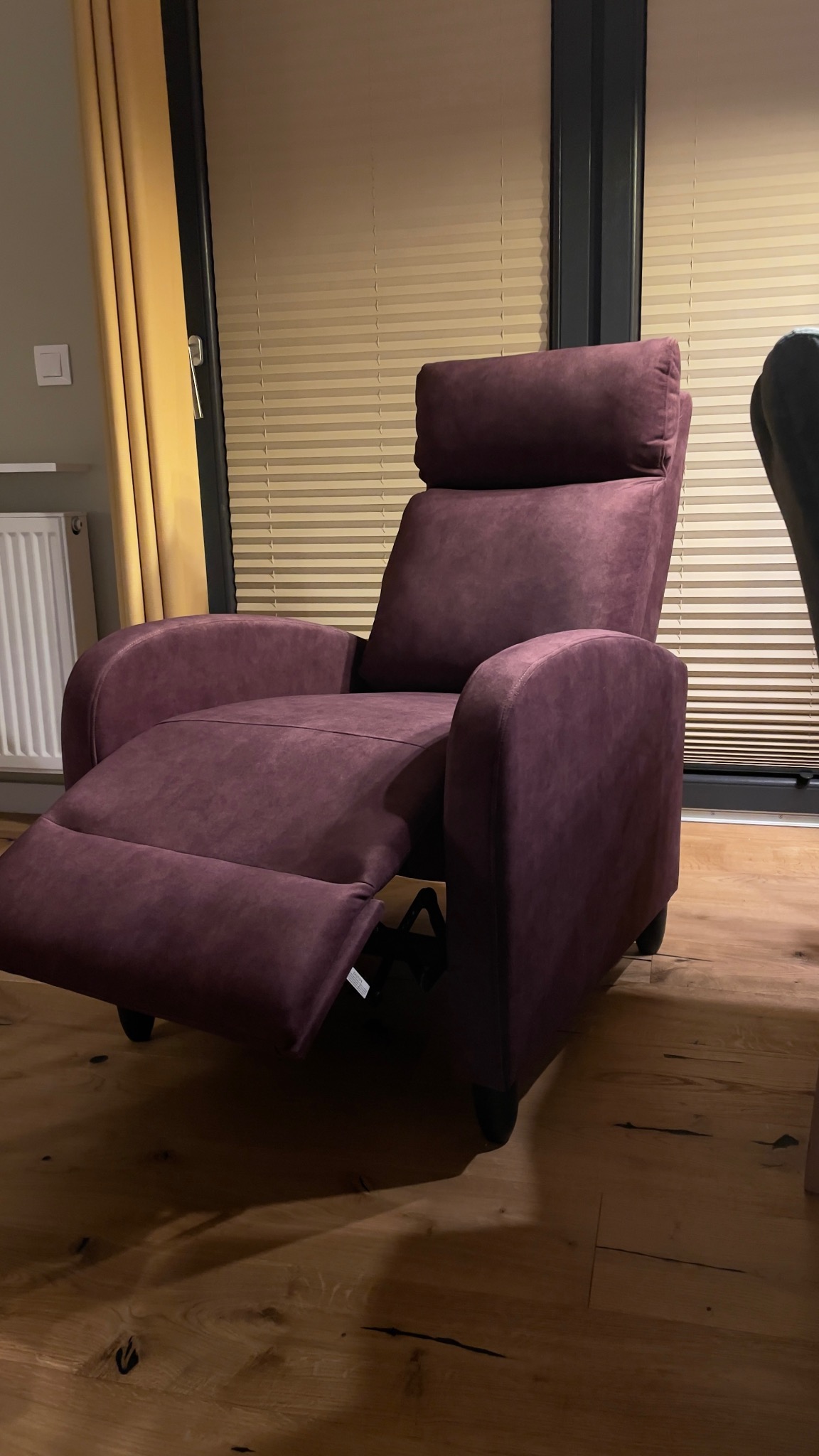 Rozłożony fotel relaksacyjny w odcieniach fioletu, z wysuwanym podnóżkiem, na drewnianej podłodze, w tle okno z żaluzjami i zasłoną.