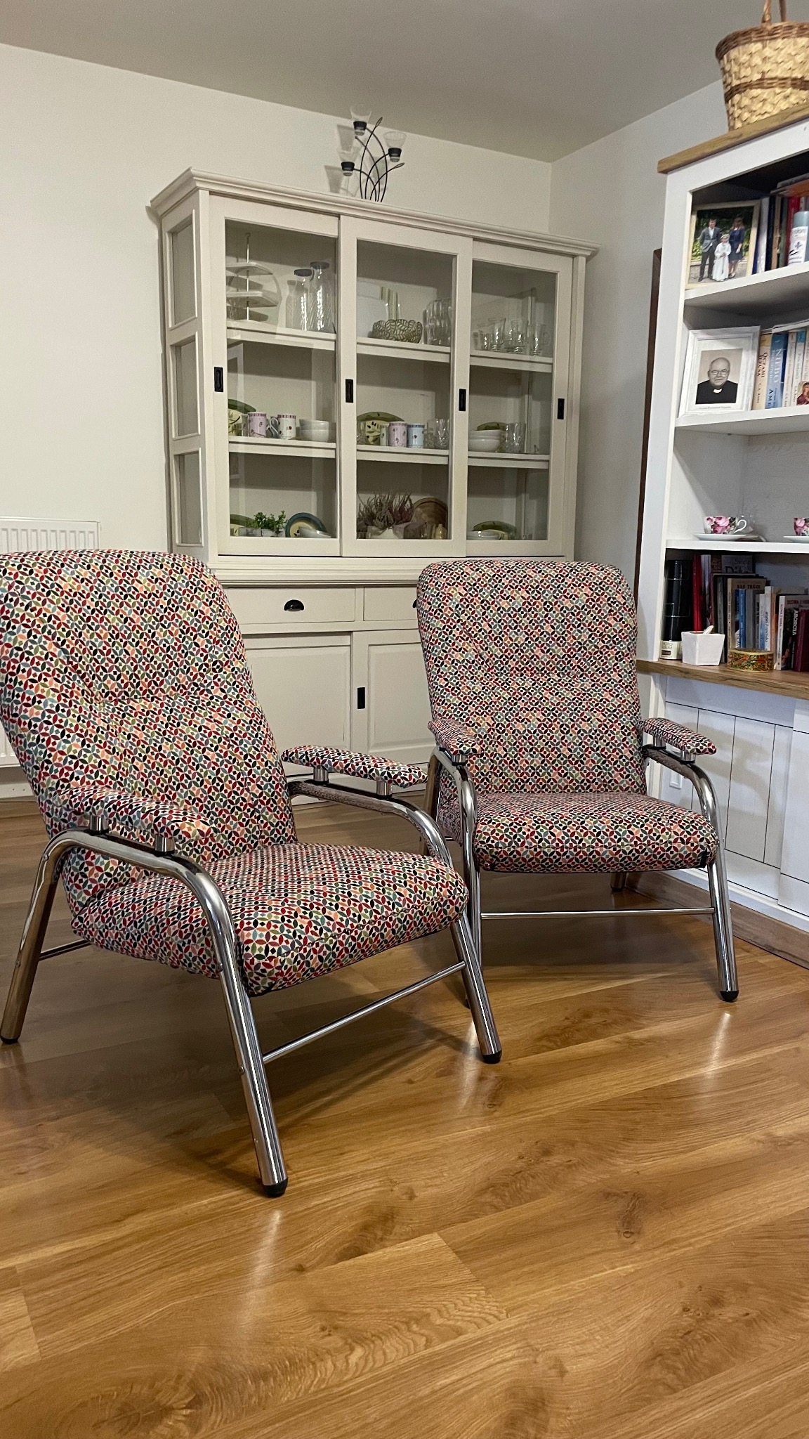 Dwa krzesła z chromowanymi ramami i tapicerką w geometryczny wzór, stojące na drewnianej podłodze na tle kredensu i regału z książkami.