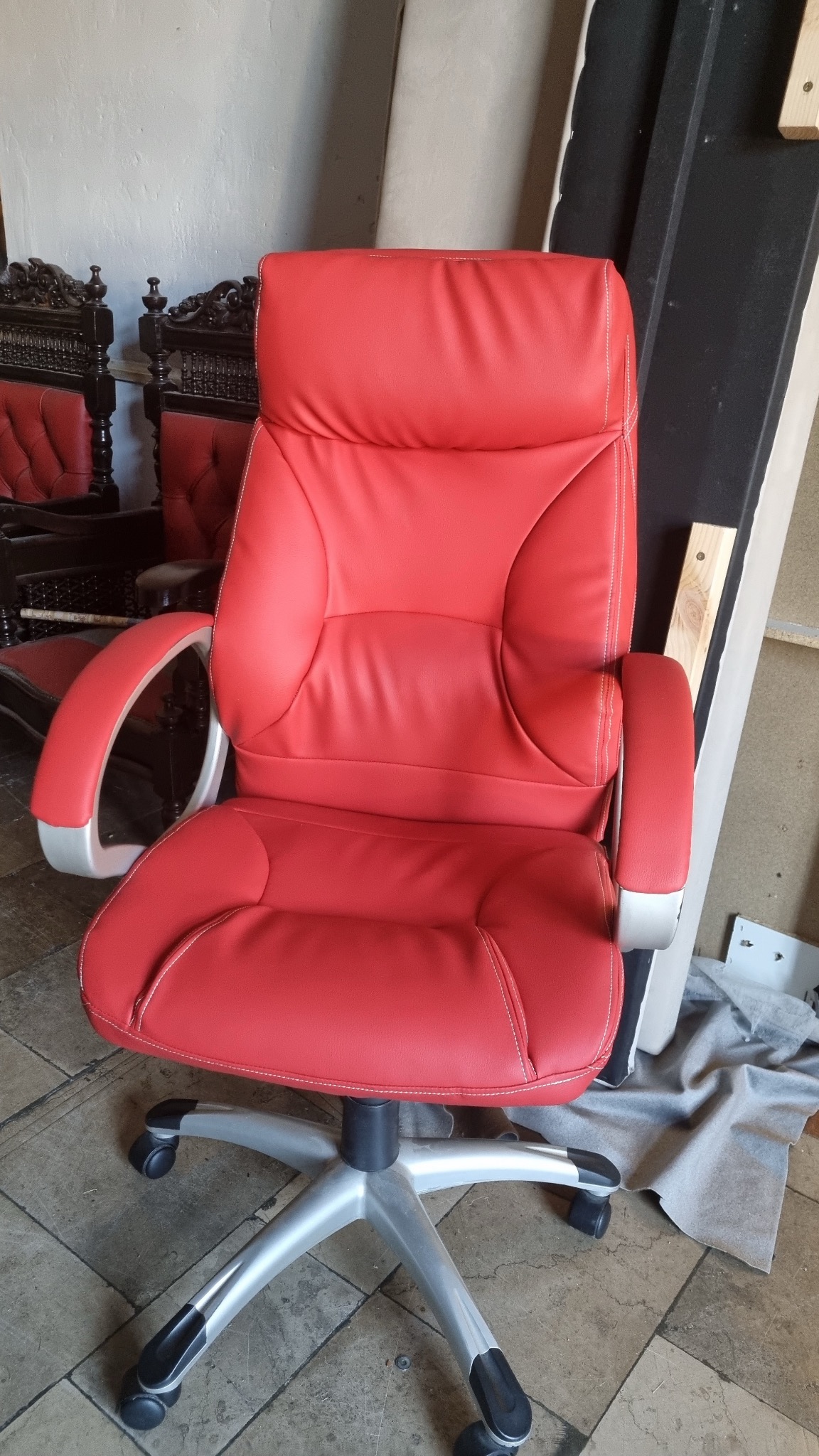 Czerwone krzesło biurowe z tapicerowanym oparciem i siedziskiem, na srebrnej podstawie z kółkami, widoczne w warsztacie obok innych mebli.