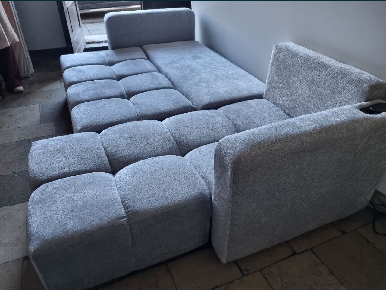 Szary, modułowy narożnik tapicerowany z widocznym uchwytem na napoje w podłokietniku, ustawiony na kamiennej posadzce.