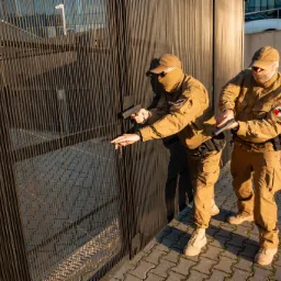 Dwóch umundurowanych strażników z bronią otwiera metalową bramę w słoneczny dzień, widoczny herb na ramieniu i budynek w tle.