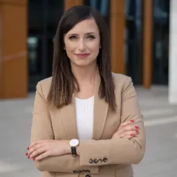 Portret kobiety w beżowym garniturze, z zegarkiem na nadgarstku, stojącej na tle nowoczesnego biurowca z przeszkloną fasadą.