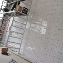 Układanie białych, kwadratowych płytek na ścianie, widok z dołu na rusztowanie z pracownikiem, wiadro z zaprawą i narzędzia na podłodze.