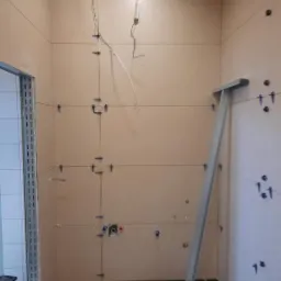 Przygotowanie ścian w łazience do układania płytek: wyznaczone linie, zamontowane uchwyty krzyżykowe, widoczne instalacje elektryczne i hydrauliczne, wspornik podtrzymujący element wyposażenia.