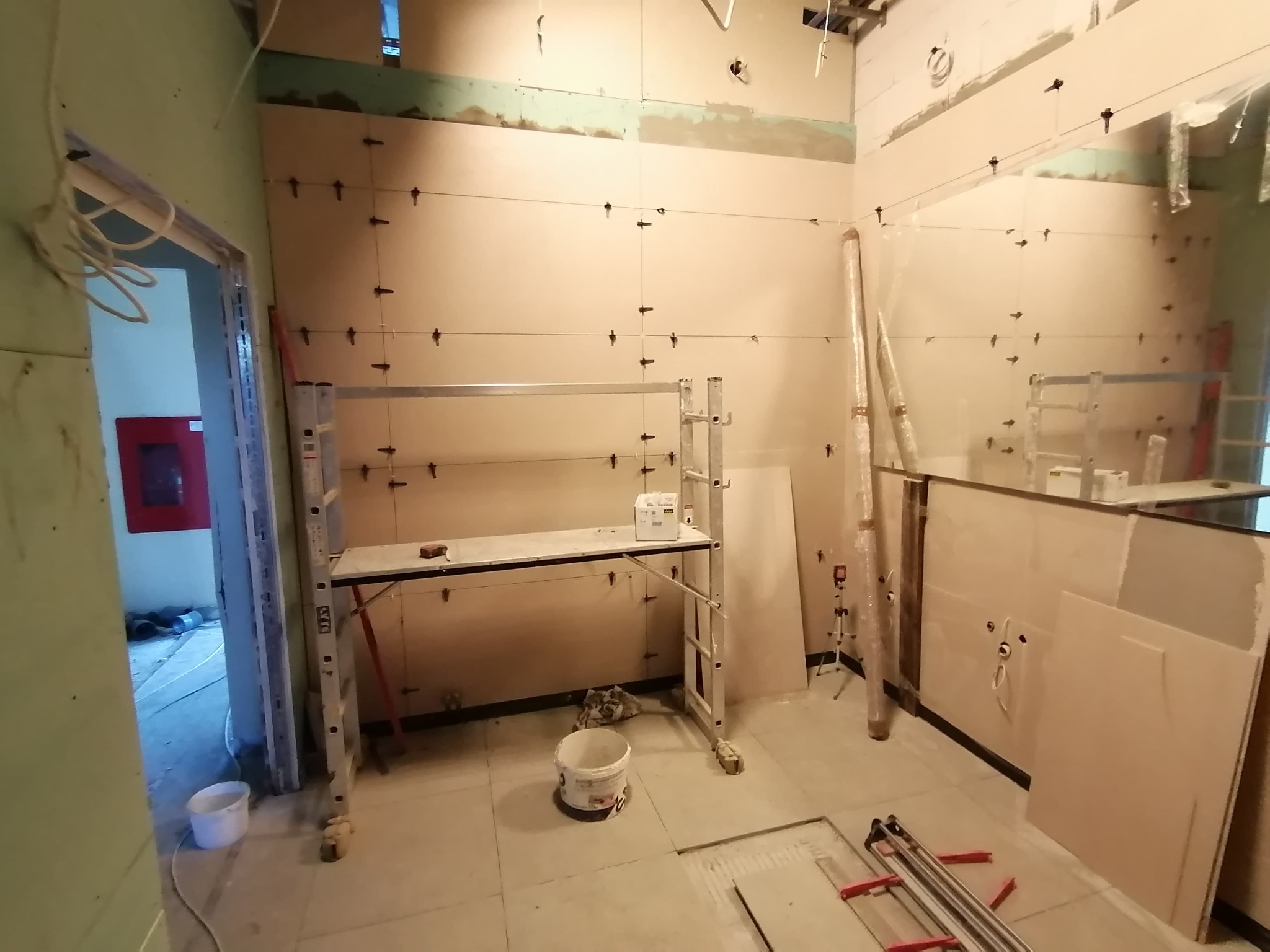 Wnętrze w trakcie układania płytek ściennych, widoczne poziomice laserowe, aluminiowy podest roboczy, wiadro z klejem i narzędzia glazurnicze.