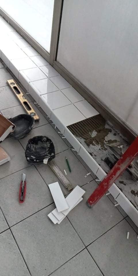 Układanie białych płytek przy oknie, widoczne narzędzia glazurnicze: poziomica, szpachelka, nóż, ołówek, zapas płytek i resztki zaprawy.