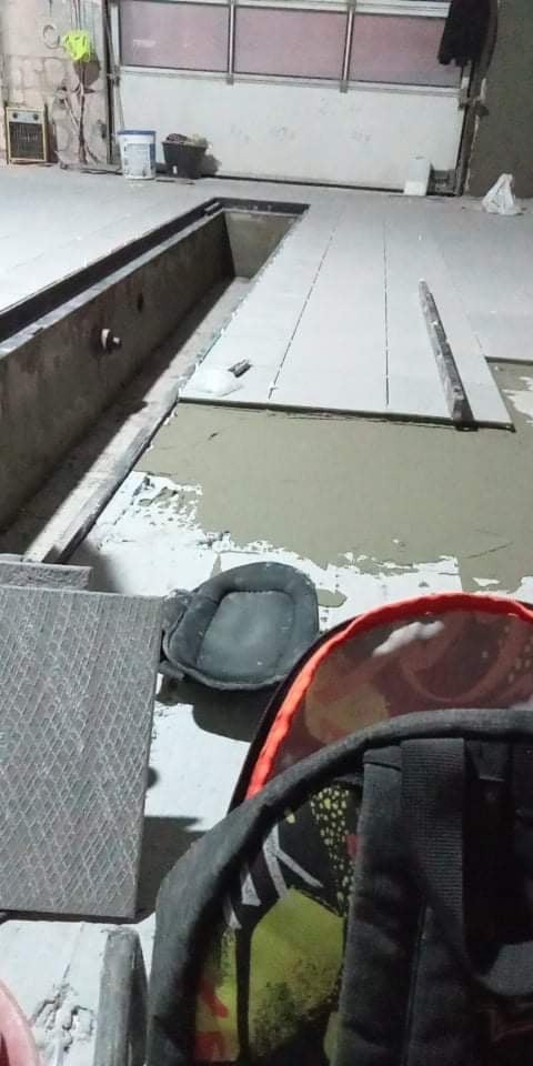 Układanie jasnych płytek podłogowych w garażu z kanałem, widoczne narzędzia i pozostałości zaprawy, fragment plecaka w pierwszym planie.