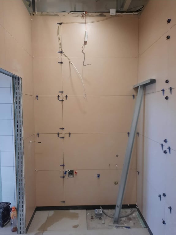 Przygotowanie ścian w łazience do układania płytek: wyznaczone linie, zamontowane uchwyty krzyżykowe, widoczne instalacje elektryczne i hydrauliczne, wspornik podtrzymujący element wyposażenia.