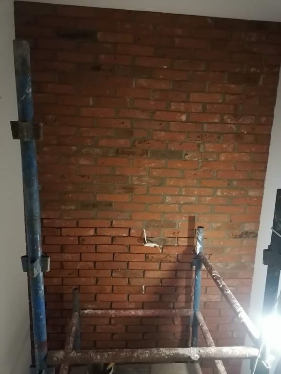Ściana z czerwonej cegły w trakcie murowania, widoczne rusztowanie i przewody elektryczne wystające ze ściany.